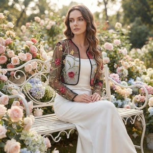 Puede incluir: Un vestido blanco largo hasta el suelo combinado con una chaqueta bolero negra transparente de manga larga con un patrón floral bordado en tonos rojo, amarillo y verde. La chaqueta tiene un borde negro a lo largo de la parte frontal abierta.