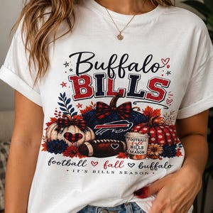 May include: A white t-shirt featuring a graphic print with the text Buffalo Bills, football, fall, buffalo, and It's Bills Season. The design includes pumpkins, a football, a coffee cup, and autumn leaves in red, blue, and black colors.