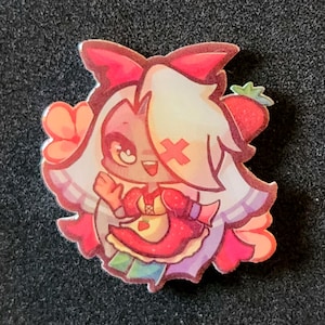 May include: A die-cut acrylic pin featuring a cartoon character with white hair, a red bow, and a red cross patch on their face. The character wears a red dress with a white apron featuring a strawberry graphic, surrounded by pink flowers and a strawberry.