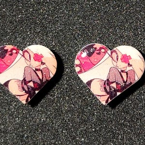 May include: Two heart-shaped pins featuring a printed illustration of a character with long blonde hair, a pink cross over one eye, and a dark outfit. The surface of the pins has a glossy, glittery finish that reflects light across the printed design.
