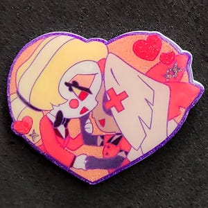 May include: A heart-shaped enamel pin featuring a cartoon illustration of two characters with blonde and white hair, wearing red and white outfits. The design includes red hearts, a purple glitter border, and a glossy finish on the surface.