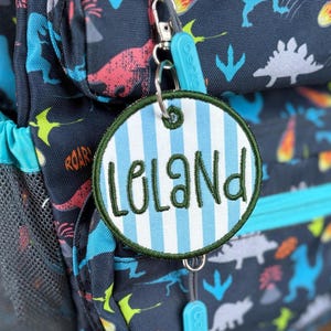 May include: A dark blue backpack featuring a colourful dinosaur pattern with a round, light blue and white striped fabric tag attached to the zip. The tag is embroidered with the name Leland in dark green cursive script and has a dark green border.