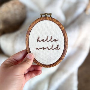 May include: An oval embroidery hoop with a faux wood grain frame containing white fabric. The words "hello world" are stitched in dark brown thread in the centre of the fabric. A small metal tightening screw is located at the top of the frame.