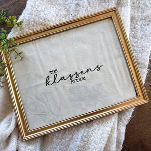 May include: A rectangular gold-toned picture frame containing a piece of off-white fabric. The fabric features black embroidered text that reads THE KLASSENS EST. 2021. The frame is positioned at an angle, showcasing its metallic finish and structure.