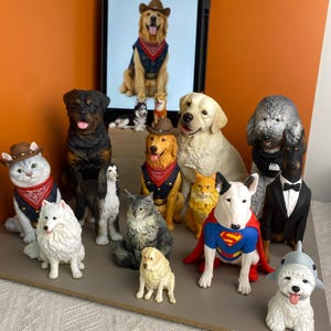 May include: A collection of various dog and cat figurines, including a bull terrier in a Superman cape, a dog in a tuxedo, and several animals wearing cowboy hats and bandanas, displayed in front of a tablet showing a golden retriever image.