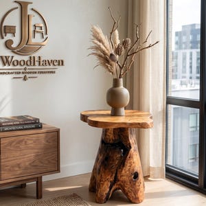 May include: A natural wood side table with a thick, organic-shaped top and a sturdy, textured tree stump base. A matte beige vase containing dried pampas grass and branches sits on the table. The wall features a bronze JLWoodHaven logo.
