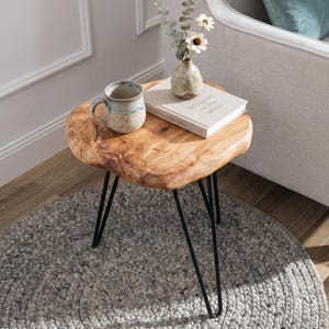 May include: A side table with a natural live-edge wood slab top and three thin black metal hairpin legs. On the table sits a speckled ceramic mug with a blue rim, a white book titled Whispers of the Wilds, and a small vase with white flowers.