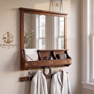 May include: A rectangular wooden wall mirror featuring an integrated shelf with three compartments, brass-toned label holders, and a lower rack with multiple hooks. The unit holds envelopes, sunglasses, a wallet, a coiled belt, and two light-colored blazers.