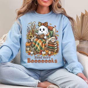 Op de afbeelding: Een lichtblauw sweatshirt met ronde hals en een afbeelding van een geest die in een stoel zit, een boek leest, gewikkeld in een plaid, omringd door pompoenen, zonnebloemen en een stapel boeken met de tekst Read more Booooooks eronder.