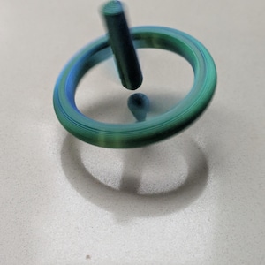 May include: A spinning top toy in motion, featuring a circular ring and a central vertical stem. The object displays a gradient of blue and green colors with visible horizontal striations throughout its structure, creating a blurred effect.