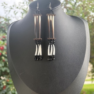 May include: Dangle earrings featuring long, thin, black and white quills arranged vertically. The design includes dark brown horizontal spacers, small black faceted beads at the top and bottom, and silver-toned metal earring hooks for attachment.