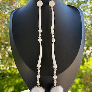 May include: Long dangle earrings featuring gold-tone hooks, white circular discs, and elongated white curved components. The design incorporates small iridescent beads, white pearls, and a soft, fluffy white pom-pom at the base of each earring.