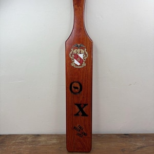 Puede incluir: Una paleta de madera larga y rectangular con un acabado marrón oscuro y un agujero en la parte superior. Presenta un escudo en la parte superior, las letras griegas Theta y Chi en negro y el texto "FALL WEEK-END 1952" impreso en negro cerca del borde inferior.