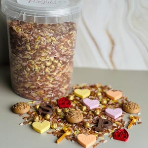 Op de afbeelding: Een doorzichtige plastic container gevuld met een mengsel van bruine, roze en gele rijstkorrels, gouden kralen en kleine decoratieve vormen, waaronder de letters S en P, hartjes en rode huisiconen. Het deksel heeft een wit label met de tekst S.P. Glitter.