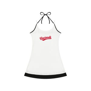 May include: A white halter-style dress featuring thin black straps that tie at the neck and a black hemline. The word Cincinnati is printed in red script across the center of the chest. The garment has a simple, sleeveless silhouette.