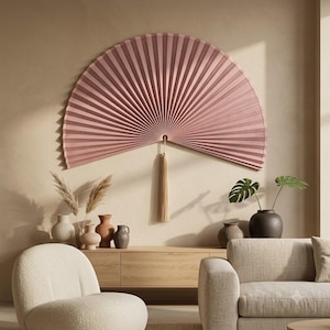 Oversized dusty rose bamboo folding wall fan with tassel styled in a cozy reading corner with wooden furniture and soft earthy decor.