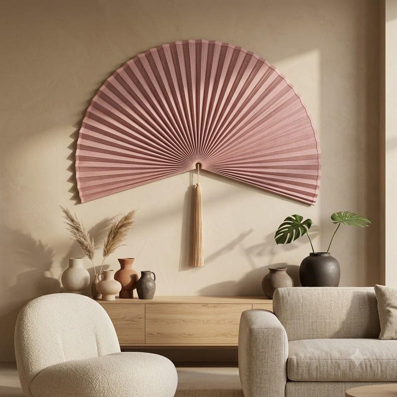 Oversized dusty rose bamboo folding wall fan with tassel styled in a cozy reading corner with wooden furniture and soft earthy decor.