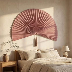 Dusty rose folding bamboo wall fan with long tassel shown on a warm beige textured wall, highlighting the full fan shape and natural rib structure.