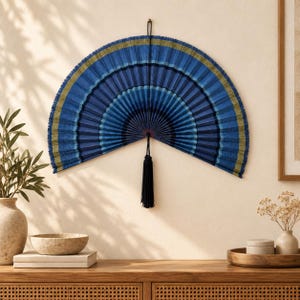 May include: A large, semi-circular wall-mounted folding fan featuring a gradient of blue and dark navy stripes with thin olive green accents. The fan is suspended by a black cord and includes a long, dark blue decorative tassel hanging from the center.