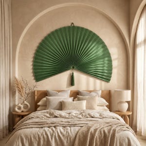 May include: A large, semi-circular, pleated dark green wall fan featuring a central hanging tassel. The fan is mounted symmetrically above a bed with neutral-toned bedding, pillows, and a textured throw blanket in a room with arched walls.