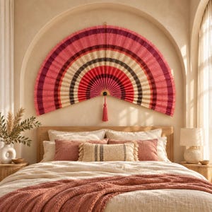 May include: A large, semi-circular wall fan featuring concentric bands of pink, red, cream, and dark brown. The fan is constructed from woven material and includes a single, long, matching pink tassel hanging from the central pivot point.