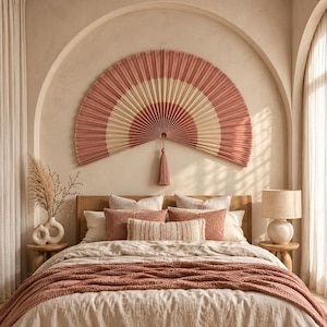 May include: A large, semi-circular wall fan featuring alternating bands of dusty rose and cream pleated fabric, centered with a matching tassel. The fan is mounted above a bed with a textured rose-colored throw and several coordinating pillows.