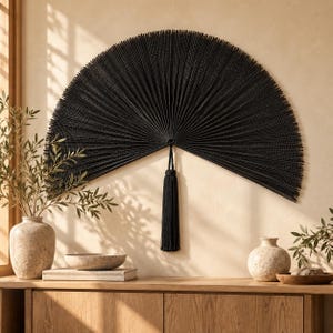 May include: A large, semi-circular black wall fan featuring a textured, pleated design that radiates from a central point. A long, matching black tassel hangs vertically from the center of the fan, adding a decorative element to the piece.