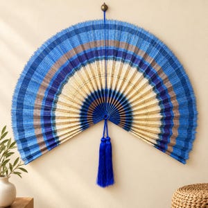 May include: A large, semi-circular wall fan featuring a pleated design with concentric bands of blue, light blue, and cream colors. The fan is suspended from a small circular hook and includes a long, vibrant blue tassel hanging from the center.