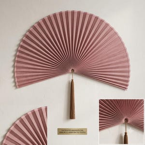 Oversized dusty rose bamboo wall fan styled above a wooden bed in a cozy neutral bedroom with soft natural light and minimalist decor.
