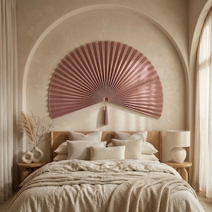 Large dusty rose bamboo wall fan with tassel displayed above a neutral upholstered bed in a warm boho bedroom with arched wall detail.