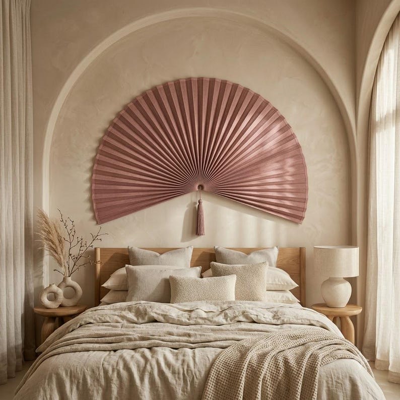 Large dusty rose bamboo wall fan with tassel displayed above a neutral upholstered bed in a warm boho bedroom with arched wall detail.