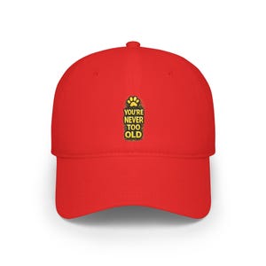 May include: A bright red baseball cap featuring a centered graphic with a yellow paw print icon above the text "YOU'RE NEVER TOO OLD" in bold yellow capital letters. The cap has a structured crown, a curved brim, and a button on the top center.