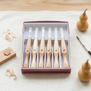 May include: A set of six wood carving tools with light-colored wooden handles and metal tips, arranged in a rectangular maroon box. Each handle features a small black character printed on the side. The tools have clear plastic protective caps.