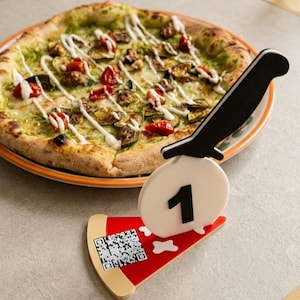 Könnte beinhalten: Eine runde Pizza mit grünem Pesto, Zucchinischeiben und roten Tomaten, beträufelt mit weißer Soße. Im Vordergrund steht ein Tischnummernhalter in Form eines Pizzastücks mit der Zahl 1, einem schwarzen Messergriff und einem QR-Code.