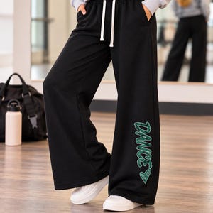 May include: Black wide-leg sweatpants featuring a white drawstring at the elastic waistband and two side pockets. The left leg displays the word "DANCE" in a green, textured, marbled font, accompanied by a small green heart outline below the text.