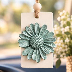 May include: A rectangular off-white hanging ornament featuring a three-dimensional sage green daisy flower attached to the center. The ornament is suspended by a brown jute twine cord accented with a single smooth, round wooden bead.