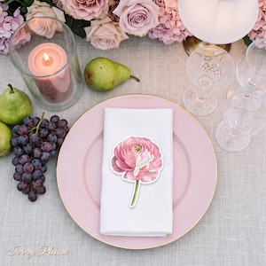 Puede incluir: Un plato llano rosa con borde dorado sostiene una servilleta blanca doblada, coronada por una tarjeta de sitio con forma de ranúnculo rosa que muestra el nombre Jennifer en letra cursiva blanca. Incluye peras verdes, uvas y una vela rosa.