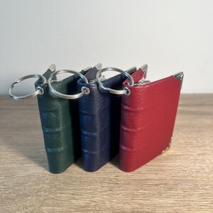 Pode incluir: Três porta-chaves em forma de livro em miniatura nas cores verde-escuro, azul-marinho e vermelho. Cada porta-chaves apresenta uma capa texturizada, um anel de metal prateado e protetores de canto decorativos de metal prateado.