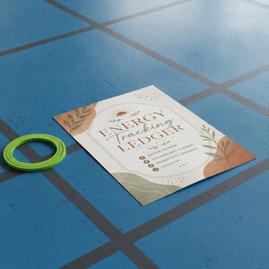 May include: A rectangular paper sheet titled ENERGY TRACKING LEDGER with a sun icon, leaf illustrations, and text listing fatigue monitor, wellness daily planner, productivity organizer, and focus log, accompanied by a small green circular ring.