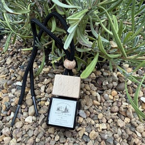 May include: A square glass car diffuser bottle with a light wood cap and a black cord. The white label features a mountain illustration and the text Pennine & Pine North Wind Fresh Linen Car Diffuser. The bottle is suspended by a black cord.