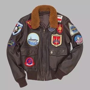 Può includere: Un giubbotto da aviatore in pelle marrone scuro con colletto in pelliccia sintetica marrone, chiusura frontale con zip e due tasche con patta. La giacca è decorata con numerosi stemmi ricamati in stile militare, tra cui una nave, testo ed emblemi.