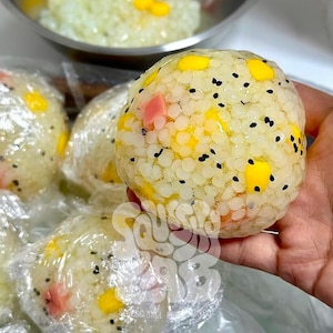 May include: A spherical rice ball wrapped in clear plastic film, featuring white rice grains mixed with yellow corn kernels, small pink ham cubes, and black sesame seeds. The texture is granular and compact, showing a colorful food composition.