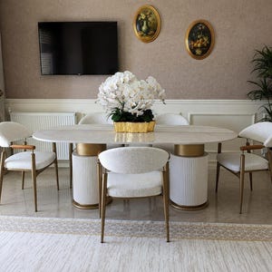 May include: An oval dining table with a white marble top and two white fluted cylindrical bases with gold accents. Four chairs with white textured upholstery, gold metal frames, and wooden armrests surround the table. A large white orchid arrangement sits in a gold rectangular planter in the centre.