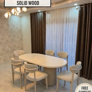 Puede incluir: Una mesa de comedor ovalada con una base cilíndrica estriada y seis sillas a juego con respaldos curvos y tapicería texturizada de color claro. El texto HANDCRAFTED DESIGN SOLID WOOD es visible en la parte superior de la imagen.
