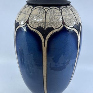 May include: A tall ceramic urn with a lid, featuring a deep blue glossy glaze and a cream-colored crackle finish. The vessel is decorated with dark, arched panels that create a repeating petal-like pattern around the body and the lid.