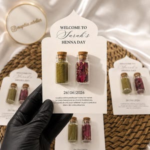 Pode incluir: Um cartão branco com borda superior decorativa apresentando o texto WELCOME TO Sarah's HENNA DAY, a data 26|06|2026 e texto em francês. Dois pequenos frascos de vidro com rolhas de cortiça estão fixados, um com pó de henna verde e outro com pétalas de rosa secas.