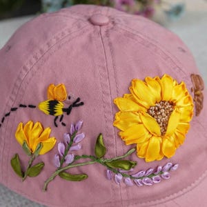 Op de afbeelding: Een oudroze baseballpet met gedetailleerd lintborduurwerk van een grote gele zonnebloem, een kleine bij met zwart-gele strepen en takjes paarse lavendel. Het ontwerp is rechtstreeks op de stoffen kroon van de pet geborduurd.