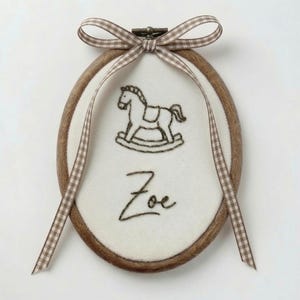 May include: An oval wooden embroidery hoop containing white fabric with a dark brown stitched rocking horse and the name Zoe embroidered in cursive script. A brown and white gingham ribbon is tied in a bow at the top of the metal tension screw.