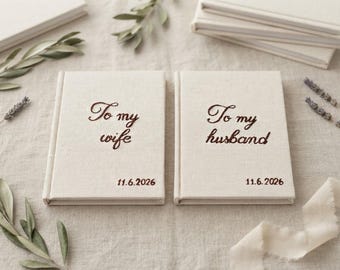 Könnte beinhalten: Zwei cremefarbene, leinengebundene Bücher mit dunkelbraunem Sticktext. Ein Buch trägt die Aufschrift To my wife, das andere To my husband. Beide Bücher zeigen das Datum 11.6.2026 in dunkelbrauner Stickerei in der unteren rechten Ecke.