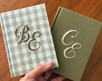 Könnte beinhalten: Zwei gebundene Bücher mit Stoffeinband. Das linke Buch hat ein salbeigrünes und weißes Vichy-Muster mit dunkelgrün gestickten Initialen BE. Das rechte Buch ist einfarbig olivgrün mit weiß gestickten Initialen CE.